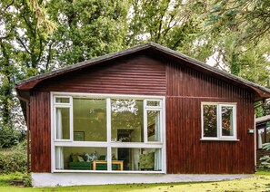 Une cabane en bois dotée de grandes fenêtres, une véranda avec un canapé vert et une petite table, le tout entouré d’une végétation luxuriante.