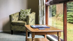 A room with a wooden table, a patterned armchair, and a window with a view of greenery.