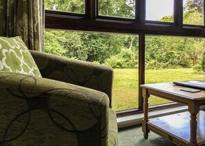 A room with a green patterned sofa, a small wooden table, and a view of a lush garden through large windows. - 1 bedroom accommodation in Ruthernbridge, Nr Bodmin (Ruthernbridge, Nr Bodmin)
