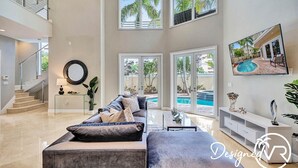 A spacious living area with a large gray sectional sofa, a coffee table, and a pool visible through the glass doors. - The Nia Tower Ultimate Beach House (Pompano Beach)