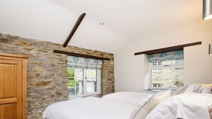 A bedroom with a stone wall, a wooden wardrobe, a bed with white bedding, and two windows with curtains. - 1 bedroom accommodation in Watergate, Looe (Watergate, Looe)