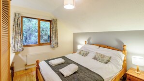 A bedroom with a wooden bed, two bedside tables, a radiator, a window with curtains, and a ceiling light. - Timber lodge set by the lake with one double and one twin. (Webbery, Bideford)