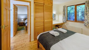A bedroom with a wooden wardrobe, a bed with towels, a desk, a window with curtains, and a radiator. - Timber lodge set by the lake with one double and one twin. (Webbery, Bideford)