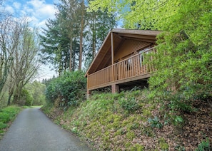 Una cabaña con terraza de madera y porche, rodeada de un bosque frondoso y un sendero pavimentado.