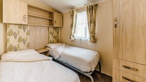 A bedroom with two beds, a wardrobe, and a window with curtains. - Modern and contemporary 12ft-wide caravan with one double and one twin. (Bleadon, Weston-Super-Mare)