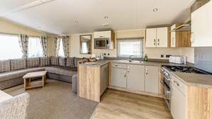 A compact kitchen with a microwave, sink, stove, and wooden cabinets. There is a seating area with a sofa and a small table. - Modern and contemporary 12ft-wide caravan with one double and one twin. (Bleadon, Weston-Super-Mare)