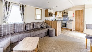 A compact living space with a sofa, a small wooden table, a kitchenette, and a dining area. - Modern and contemporary 12ft-wide caravan with one double and one twin. (Bleadon, Weston-Super-Mare)