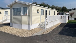 A single-story mobile home with a front porch, a small staircase, and a chimney. - Modern and contemporary 12ft-wide caravan with one double and one twin. (Bleadon, Weston-Super-Mare)