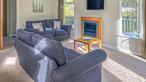 A living room with a sofa, a TV on a stand, and a window with a view of greenery. - Decadent and spacious contemporary timber lodge. (Whitfield, Hexham)