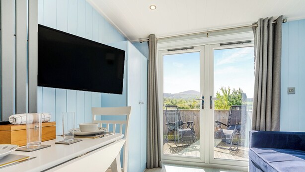 Une pièce dotée d’un téléviseur fixé au mur, d’une table à manger avec des chaises, d’un canapé et d’une porte-fenêtre coulissante donnant sur une terrasse extérieure avec vue sur la verdure et les collines.