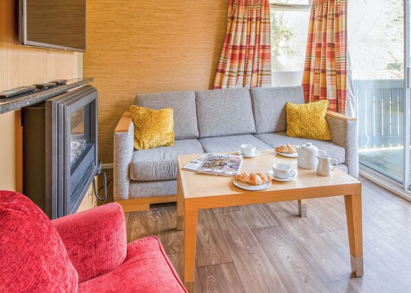 Een woonkamer met een grijze bank, gele kussens, een houten salontafel met croissants en kopjes, een televisie en een rode fauteuil.