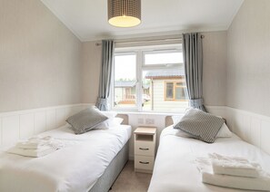 A bedroom with two beds, a window with curtains, a bedside table, and a lamp. - 2 bedroom accommodation in Hunstanton (Hunstanton)