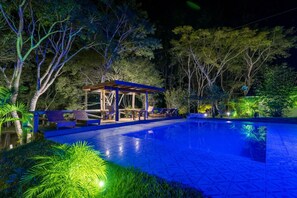Un área de piscina nocturna con zona de estar techada y sillones reclinables.