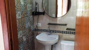 Um banheiro com parede azulejada, pia com coluna, vaso sanitário e espelho redondo.