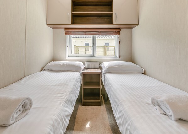 A small, compact room with two single beds, a window with blinds, a radiator, and a wooden staircase.