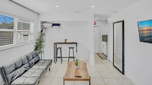 A modern, well-lit room with a leather sofa, a wooden table with a remote control, a small bar area, and a large window with blinds.