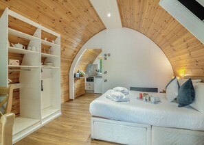 Een gezellige, compacte slaapkamer met een gebogen plafond, een bed met kussens en een dienblad, een witte garderobe en een kitchenette op de achtergrond.