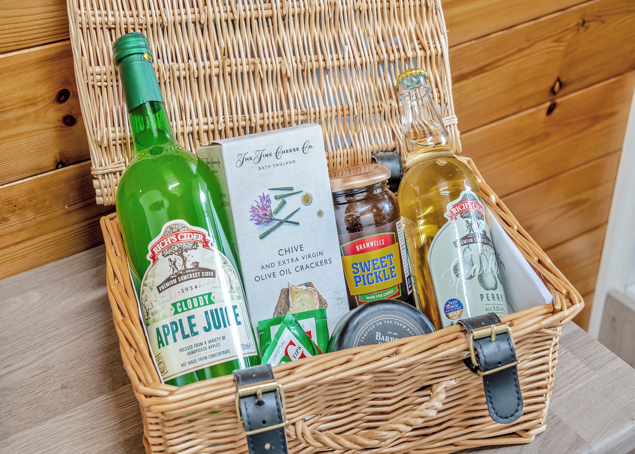 A wicker basket containing a green bottle of apple juice, a box of chive and extra virgin olive oil crackers, a jar of sweet pickle, a bottle of perry, and a tin of bars.
