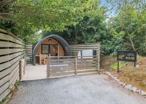 Een houten boogpoort die leidt naar een gezellige, gedeeltelijk open constructie met een bankje en een tafel, omringd door groen.