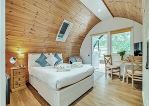 A bedroom with a wooden ceiling, a bed with pillows, a bedside table with a lamp, and a view of the outdoors.