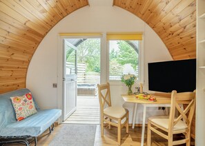 A cozy room with a wooden ceiling, a sofa with a floral pillow, a dining table with chairs, and a view of a garden through a door.