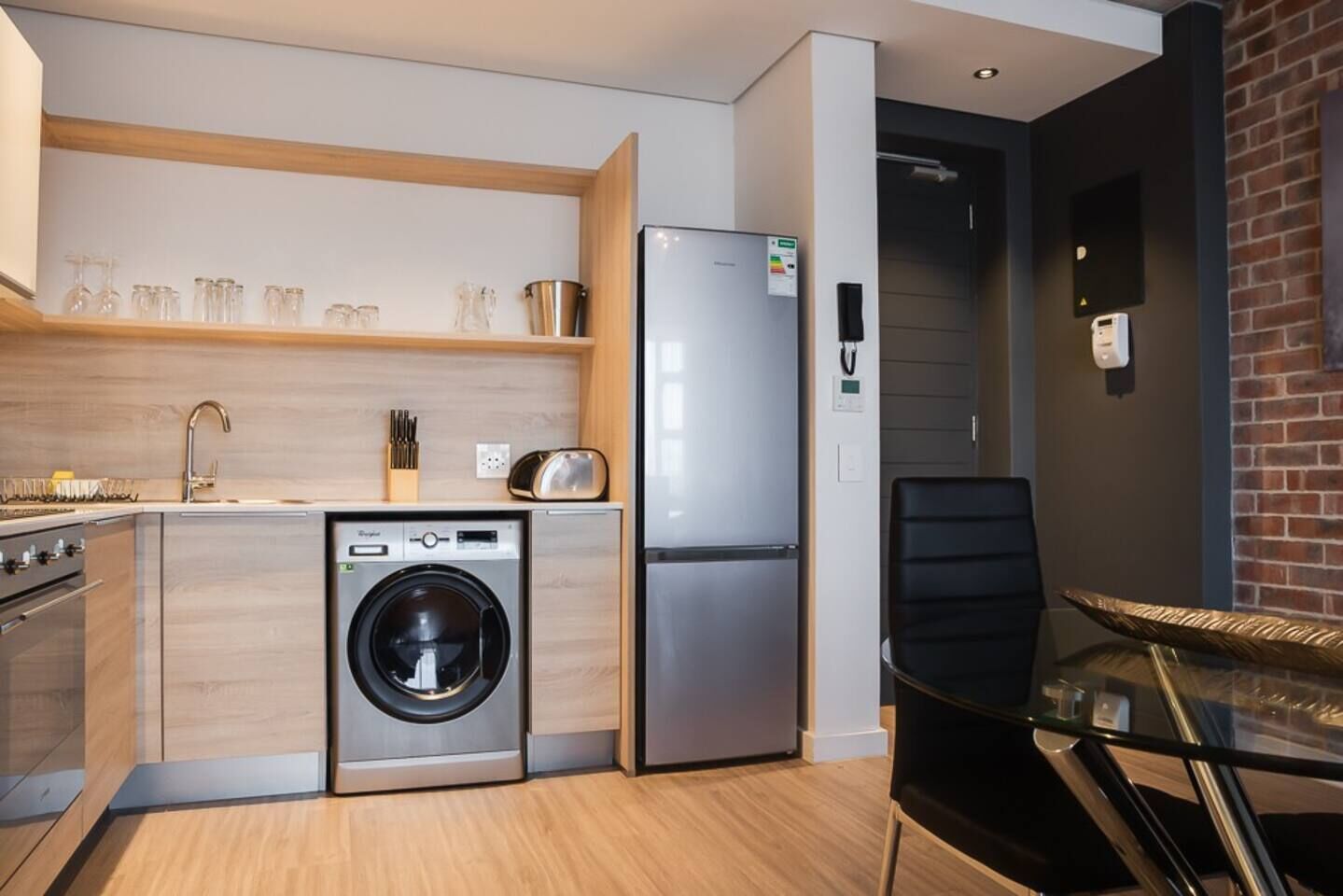 Cozinha compacta com máquina de lavar, geladeira e área de refeições com mesa de vidro.