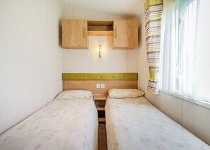 A small room with two beds, wooden headboards, a light fixture above the beds, and a window with curtains.