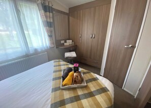 A hotel room with a bed, a checkered bedspread, a tray with towels and a cup, a wooden wardrobe, and a window with curtains.