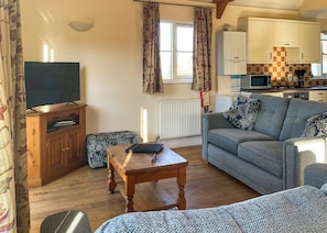 A living room with a sofa, a wooden table, a television, and a kitchen area with cabinets and appliances. - 2 bedroom accommodation in Palgrave, Diss (Palgrave, Diss)