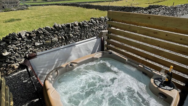 A hot tub with a stone wall and wooden bench, set against a rural landscape with sheep and a stone building. - 1 bedroom accommodation in Hawes, (Hawes,)
