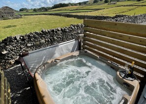 A hot tub with a stone wall and wooden bench, set against a rural landscape with sheep and a stone building.