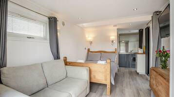 Une chambre à coucher moderne avec un lit en bois, un canapé gris, une petite table et, au fond, un coin cuisine.