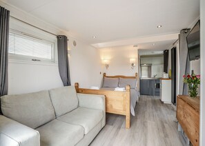 A modern bedroom with a wooden bed, a grey sofa, a small table, and a kitchenette in the background.
