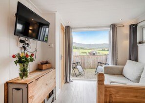 A room with a TV, a wooden desk, a sofa, and a balcony with a view of the countryside.