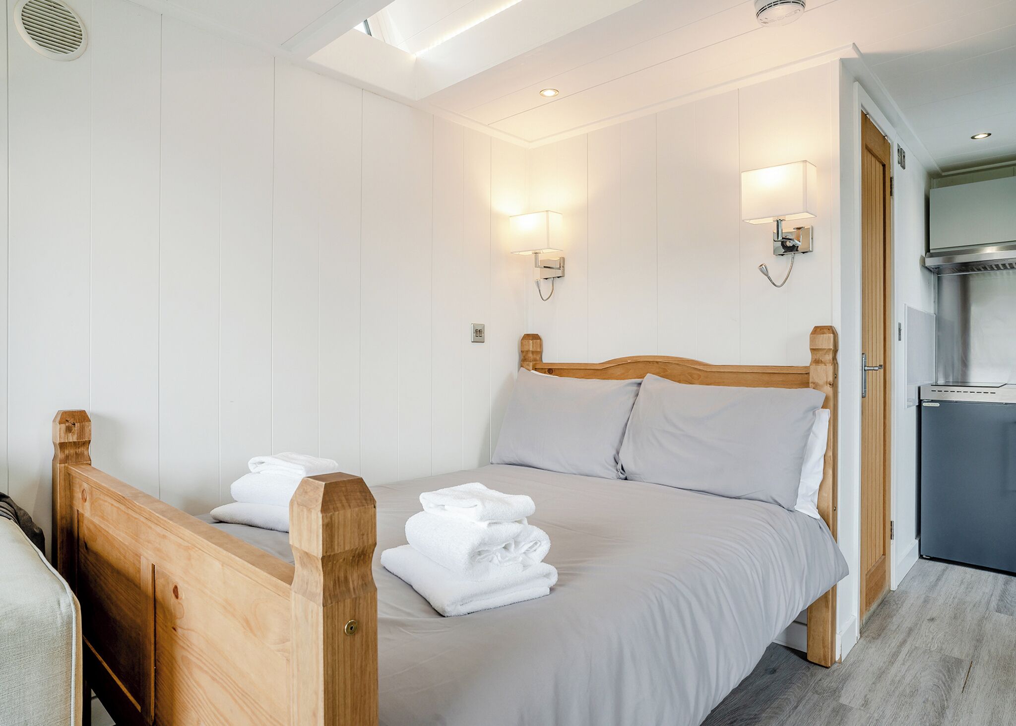 Une chambre avec un lit en bois, du linge de lit blanc, deux lampes fixées au mur, un bureau intégré et un coin cuisine.