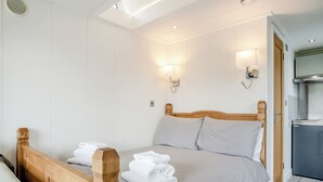 Un dormitori amb llit de fusta, roba de llit blanca, dues làmpades penjades a la paret, escriptori integrat i una cuineta.
