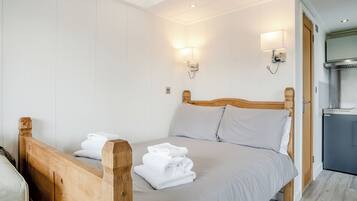 Une chambre avec un lit en bois, du linge de lit blanc, deux lampes fixées au mur, un bureau intégré et un coin cuisine.