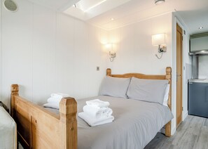 A bedroom with a wooden bed, white linens, two wall-mounted lights, a built-in desk, and a kitchenette.
