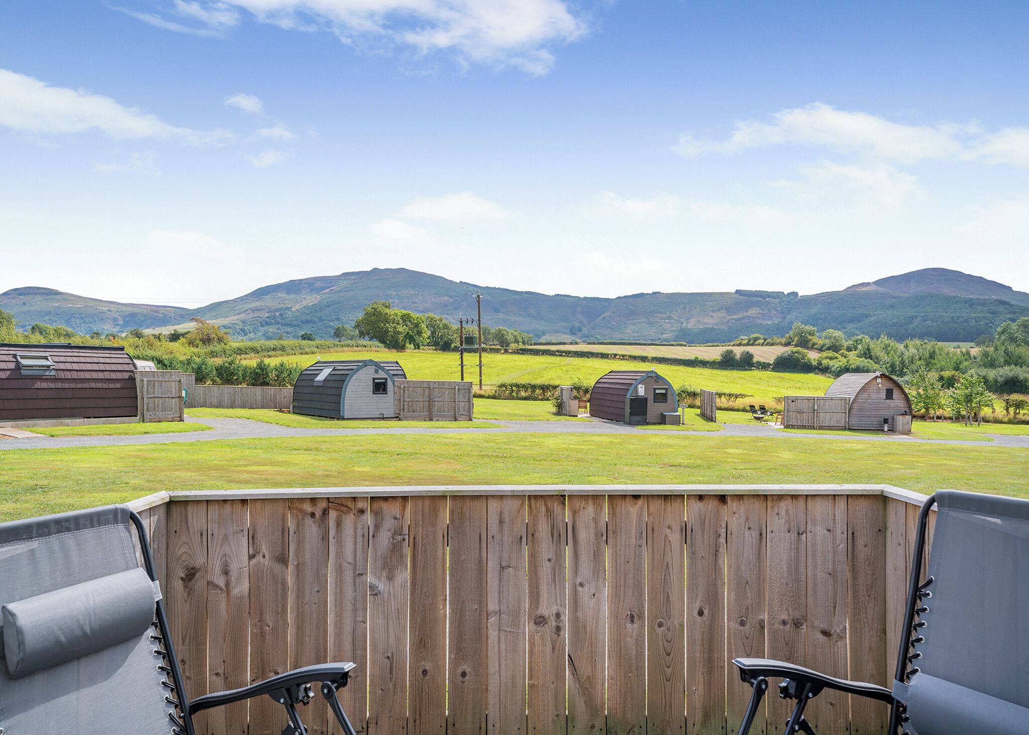 Une terrasse avec deux chaises, une clôture en bois et plusieurs petits abris de jardin, le tout situé dans un cadre rural avec des collines en arrière-plan.