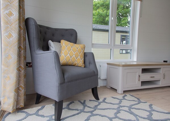 Un fauteuil gris agrémenté d’un coussin à motifs, une armoire blanche et un tapis à motifs dans une pièce dotée de grandes fenêtres.