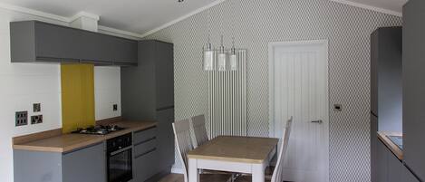 A compact kitchen with a dining area, grey cabinetry, a wooden table, and a patterned wallpaper.