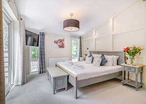 A bedroom with a large bed, bedside tables, a bench, a window with curtains, a radiator, and a wall-mounted artwork.