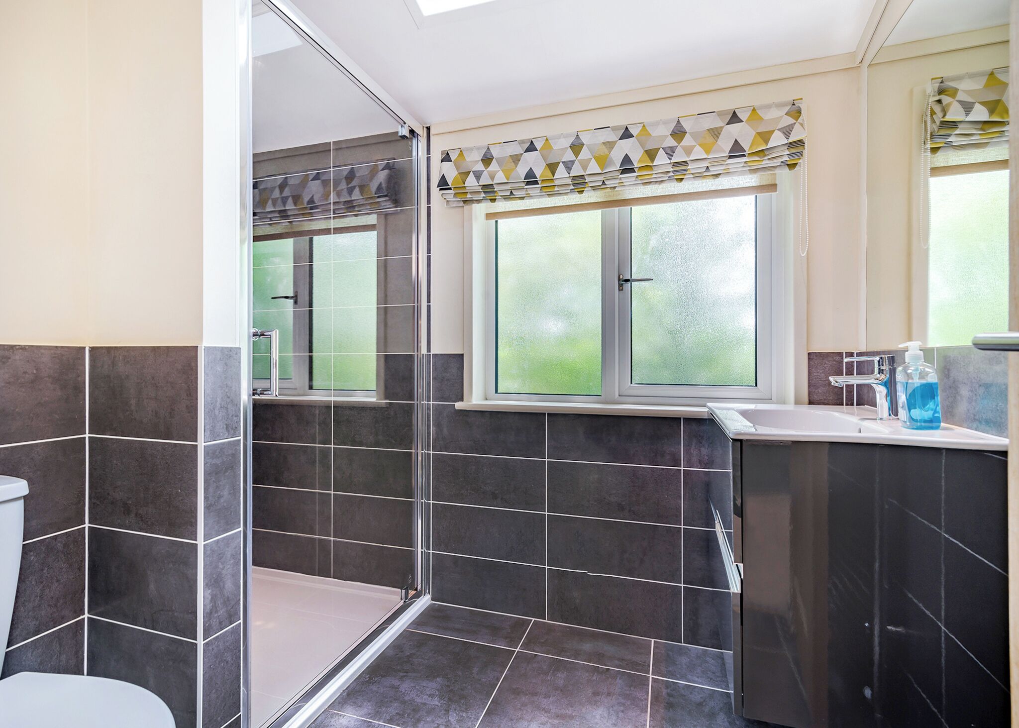 Une salle de bain équipée d’une douche, de toilettes, d’un lavabo et d’une fenêtre agrémentée d’un rideau à motifs géométriques.