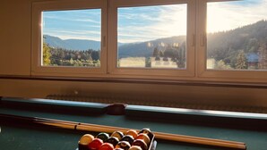 Une pièce avec une table de billard, trois fenêtres donnant sur une forêt et des montagnes, et des lampes suspendues.