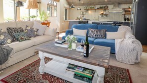 A spacious living room with a blue sofa, a white sofa, and a wooden coffee table with books and a bottle. There are large windows and a kitchen in the background. - The Owl's Shack (Kendal)