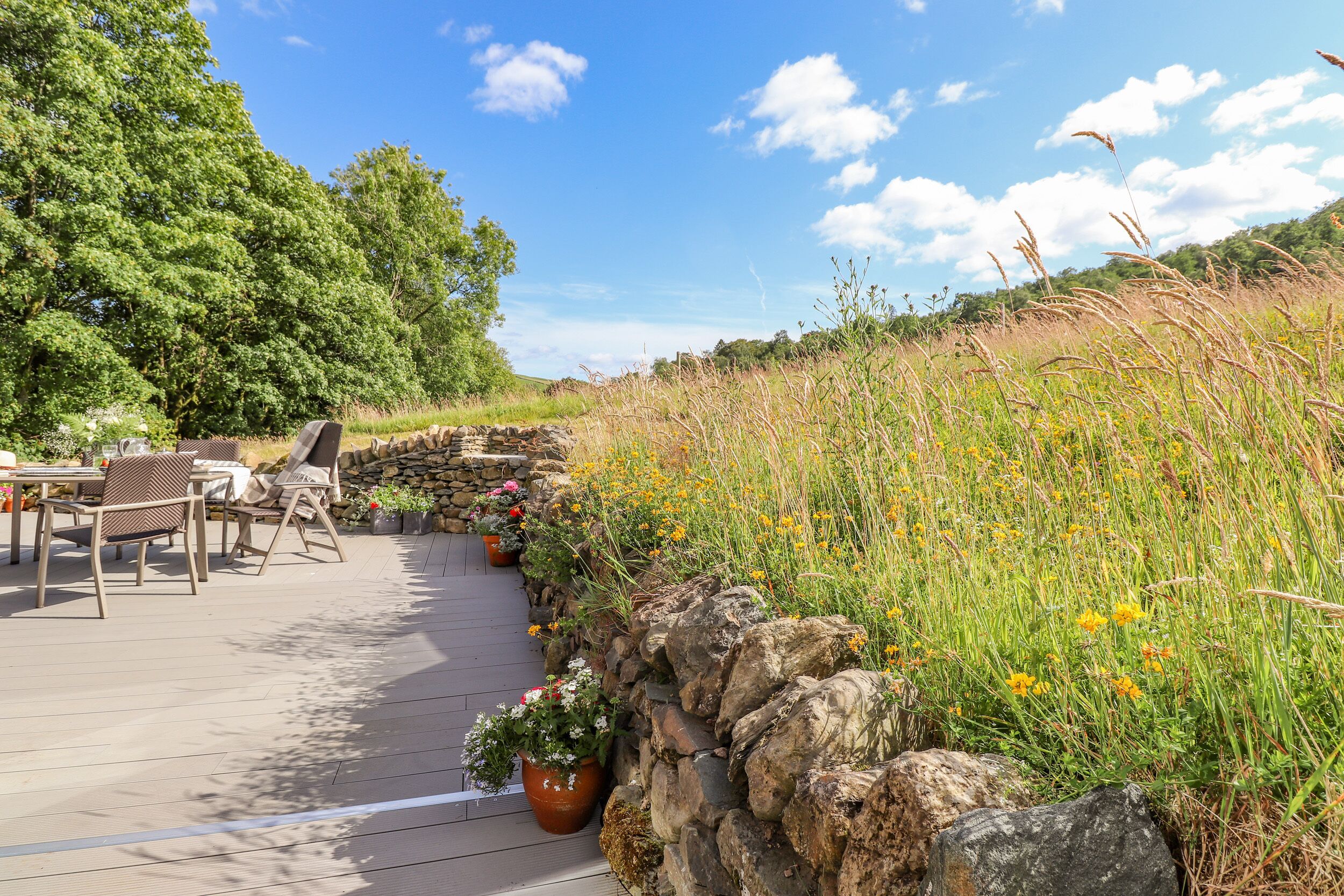 A wooden deck with outdoor furniture, a stone wall, and a grassy area with wildflowers.