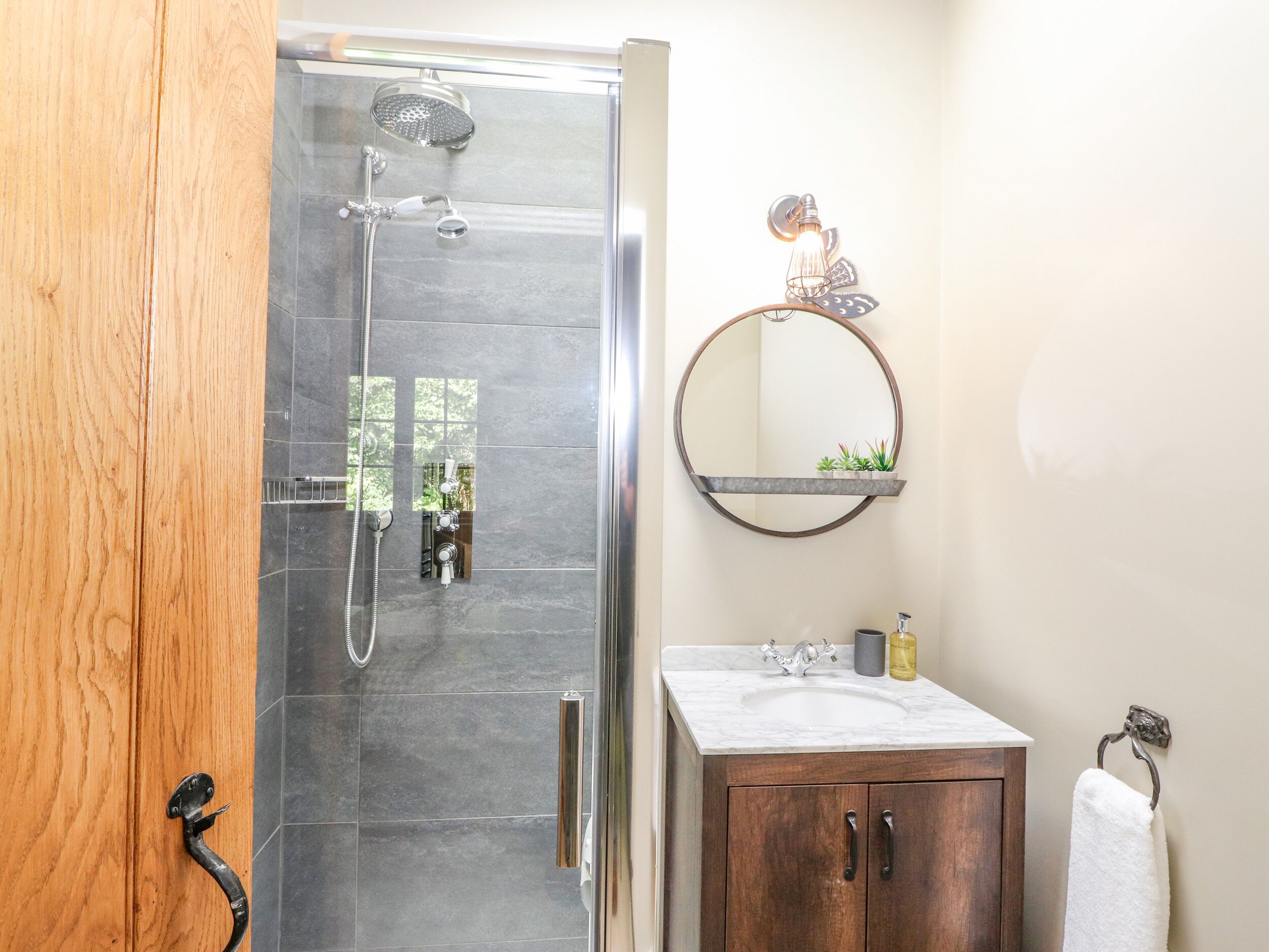 A bathroom with a glass shower enclosure, a wooden door, a marble sink, and a round mirror with a shelf above it.