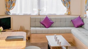 A modern room with a sofa, a coffee table, a TV, and a window with curtains. - New for 2019. Contemporary caravan with one double and two twin. (Cargill)