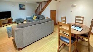 A living room with a grey sofa, wooden dining table with chairs, a TV on a wooden stand, and a staircase. - Cornish end terraced cottage with one double and one twin. (Tintagel)