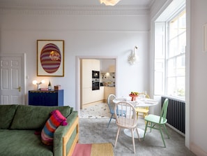 A modern living room with a green sofa, colorful pillows, a dining table with chairs, and a kitchen in the background.